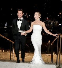 a man and woman dressed in formal wear walking down the stairs at night with their arms around each other