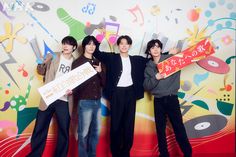 four young men holding up signs in front of a colorful wall with music notes on it