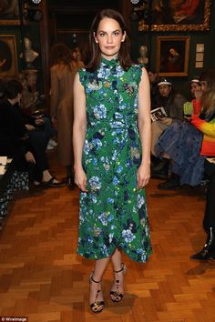 a woman in a green floral dress standing on a wooden floor with other people behind her