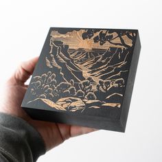 a hand holding up a wooden box with an image of mountains and trees on it