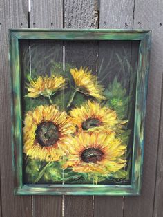 a painting of sunflowers in a green frame hanging on a wooden fence outside