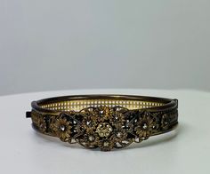 Antique gilt hinged bangle bracelet with floral bouquet and marcasite details, 1940s vintage, dainty details. A geometric like base surrounds the wrist and the front secton has a lovely floral motif with bouquets of sweet flowers. Each of the flowers has a center of a faceted marcasite stone. The clasp and hinge work well, you may need to guide the clasp in gently but it is secure once latched. The color of the piece is an antiqued gold tone or gilt color. There is no makers mark. If you are concerned about fit, we recommend comparing our measurements to a similar item in your collection that you know fits you well.  Specs at a glance Width: 3/4 inches at widest point Interior Diameter: 2.25 Interior Circumference: 6.5 inches We have much more in store and in our showroom than online. Plea Elegant Bangle With Antique Finish, Vintage Rose Cut Diamond Bracelet, Vintage Hinged Bracelets For Wedding, Vintage Rose Cut Diamond Bracelet Gift, Victorian Brass Wedding Bracelets, Victorian Wedding Bracelets In Antique Gold, Vintage Bangle With Intricate Design, Vintage Wedding Bracelets With Antique Finish, Victorian Antique Gold Bracelets For Wedding