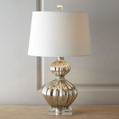 a lamp on a table in front of a window with a glass base and white lampshade