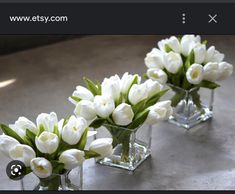 three clear vases filled with white tulips on top of a gray table