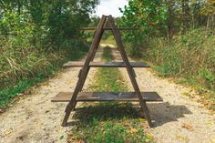 a wooden bench sitting in the middle of a dirt road next to grass and trees