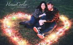 a man and woman sitting on the ground in front of a heart made out of lights