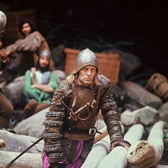 a man dressed in medieval armor standing next to another man on a rock covered beach