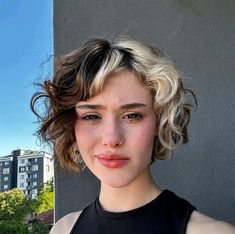 a close up of a person wearing a black shirt and blonde hair with buildings in the background