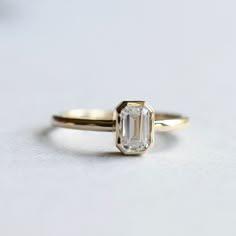 an emerald cut diamond sits on top of a yellow gold engagement ring with a thin band