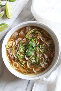 a white bowl filled with noodles and meat on top of a marble counter next to sliced limes