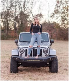 a woman standing on the hood of a jeep in a field with trees behind her