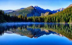 a lake surrounded by mountains and trees with water in the foreground on a sunny day