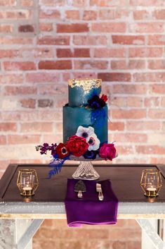 a blue and purple wedding cake with red flowers on the top is sitting on a table in front of a brick wall