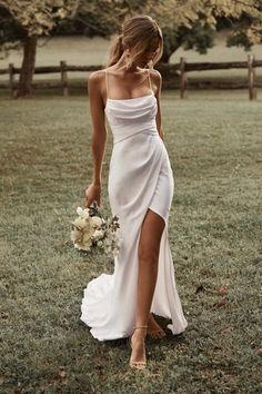 a woman in a white dress holding a bouquet and walking through the grass with her hand on her hip