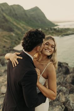 a man and woman embracing each other on the rocks by the ocean with mountains in the background