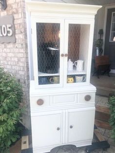 a white china cabinet sitting on top of a brick floor next to bushes and potted plants