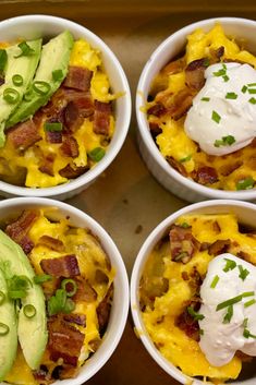 four white bowls filled with breakfast food on top of a wooden table next to an avocado and sour cream