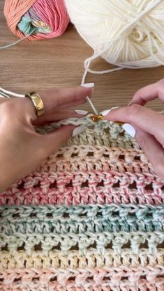 two hands crocheting together with yarn on a table next to balls of yarn