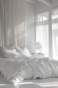 an unmade bed in a white bedroom with sheer curtains and sun shining through the window