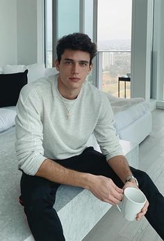 a young man sitting on the edge of a bed holding a cup in his hand