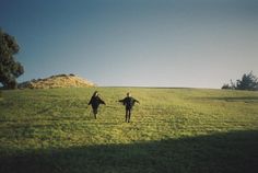 two people walking across a lush green field next to a tree on top of a hill