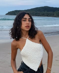 a woman standing on top of a sandy beach next to the ocean with her hands in her pockets