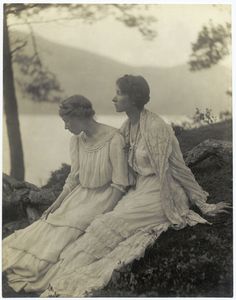 two women sitting next to each other on top of a hill near the water and trees