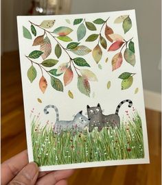 a hand holding up a card with two cats in the grass and leaves on it