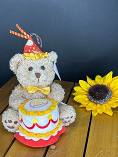 a brown teddy bear sitting next to a sunflower on a wooden table with a birthday cake