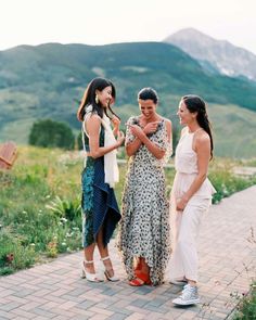 three women standing next to each other on a brick walkway with mountains in the background