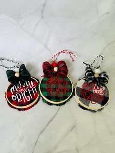 three christmas ornaments are hanging on a white tablecloth with black and red ribbon, one is saying merry bright the other says ready to decorate