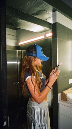 a woman standing in front of a mirror looking at her cell phone while wearing a blue hat