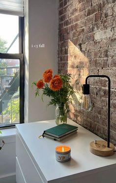 an orange flower in a vase next to a book and candle on a white table