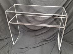 a clear acrylic table with two legs and a shelf on one side, against a gray background