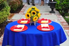 a blue table topped with red plates covered in yellow and white napkins next to a vase filled with sunflowers