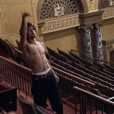 a shirtless man standing in an auditorium holding his arms up to the ceiling and looking up