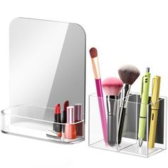 an assortment of cosmetics and makeup brushes in a clear holder next to a mirror on a white background