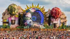 a large crowd of people standing in front of an outdoor stage with giant sculptures on it's sides