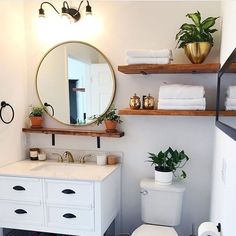 a white toilet sitting next to a bathroom sink under a round mirror and wooden shelves