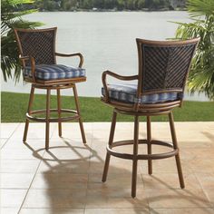 two wicker barstools with blue cushions sit in front of a lake and palm trees