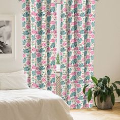 a white bed sitting next to a window covered in pink and blue curtains on top of a wooden floor