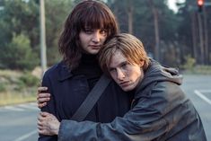 two people standing next to each other in the middle of an empty street with trees behind them