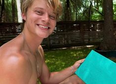 a shirtless young man sitting on the grass holding a blue book in his hands