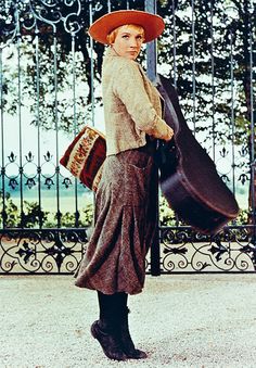 a woman wearing a hat and dress holding a suitcase in front of an iron gate