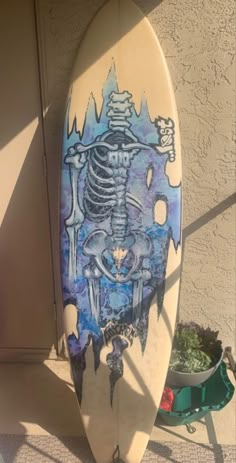 a blue and white surfboard with a skeleton painted on it's side next to a potted plant