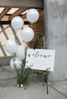 a welcome sign with white balloons and greenery in front of a concrete building on the sidewalk