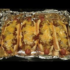 four hotdogs covered in cheese and chili sitting on tin foil lined up against each other