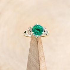 an emerald and diamond ring sitting on top of a wooden stand