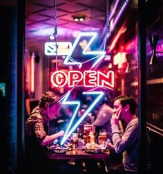 two men sitting at a table in front of a neon sign that reads open with a lightning