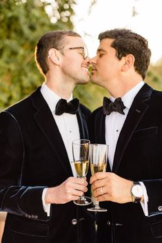 two men in tuxedos are kissing each other with champagne glasses on their hands